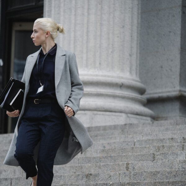 Serious female lawyer in elegant outfit holding folders with documents while going downstairs near stone state building and looking away